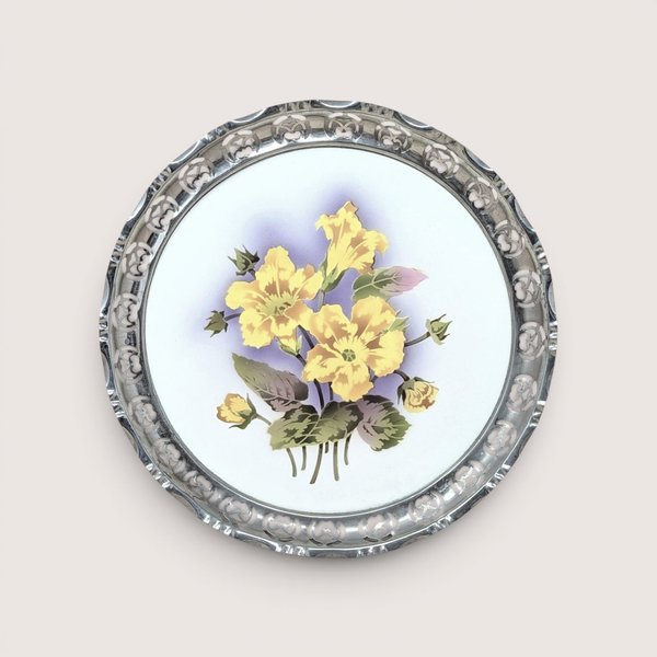 A round porcelain plaque with painted yellow flowers and a pierced silver rim, centered on a beige background.