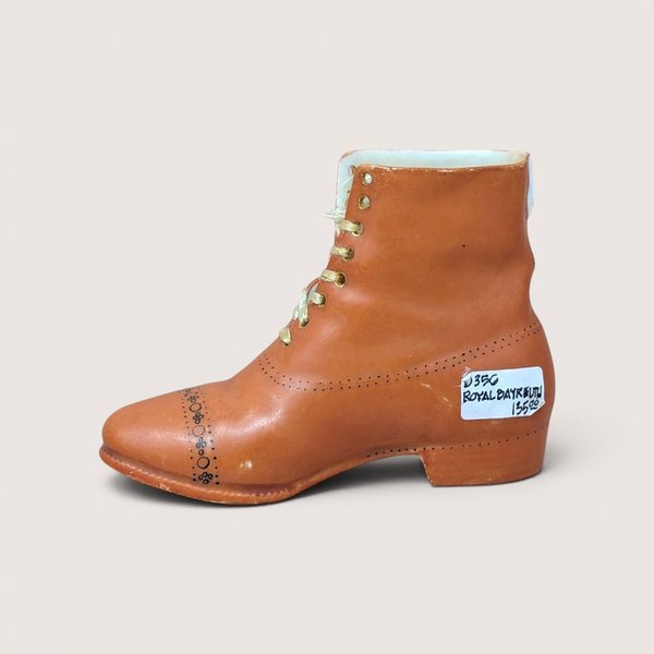 A vintage brown porcelain high-top boot figurine with a handwritten tag, centered on a beige background.