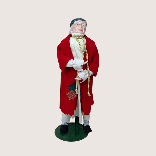 A 1987 vintage Department 56 Ebenezer Scrooge doll on a tan textured background. The nineteen inch tall figure features a detailed porcelain face with wire glasses, dressed in a red velvet night robe and holding a gold rope cord on a green metal stand.
