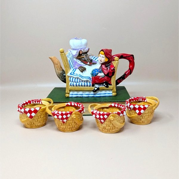 A colorful Dept 56 ceramic teapot set showing Little Red Riding Hood sitting by the Big Bad Wolf in a bed, accompanied by four small tea cups shaped like wicker picnic baskets