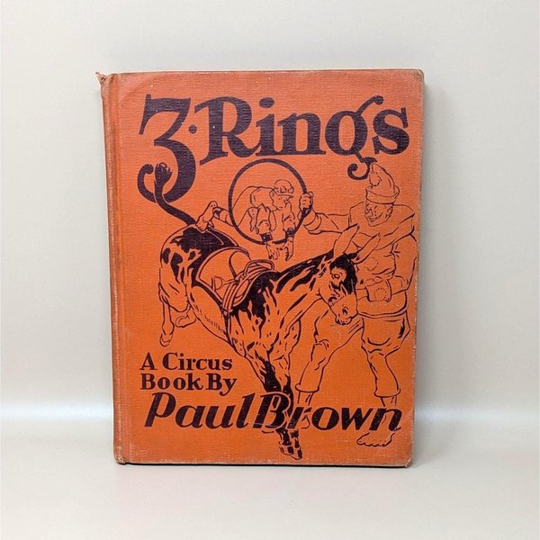 A vintage 1938 hardcover book titled 3 Rings A Circus Book by Paul Brown with an orange cloth cover featuring a black line drawing of a circus clown and donkey