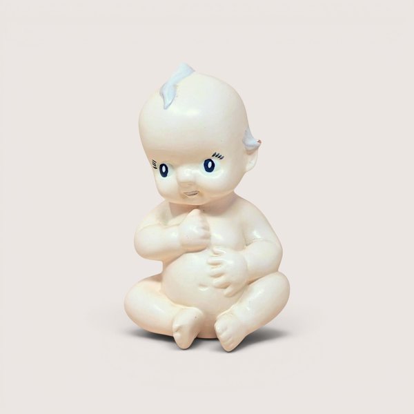 A vintage ceramic Kewpie-style baby figurine sitting down, centered on a beige background.