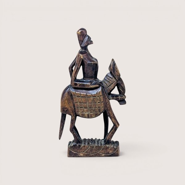 A carved dark wood figurine of a person riding a donkey or mule, centered on a beige background.