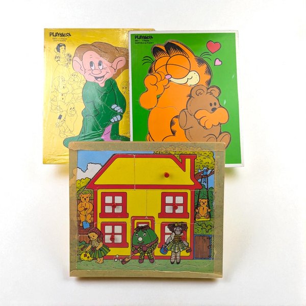 A set of three vintage wooden puzzles: one green puzzle with Garfield hugging a bear, one yellow puzzle with the dwarf Dopey, and one square puzzle of a house with small red pegs.