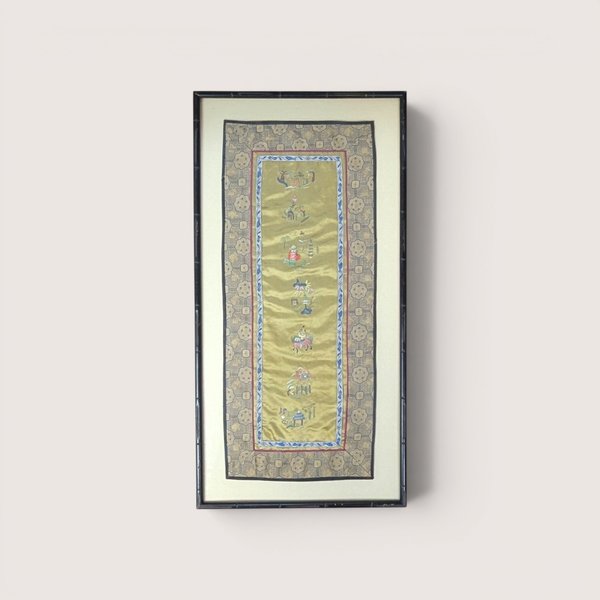 Framed vertical Chinese silk embroidery panel with intricate needlework figurines and a patterned border on a neutral wall