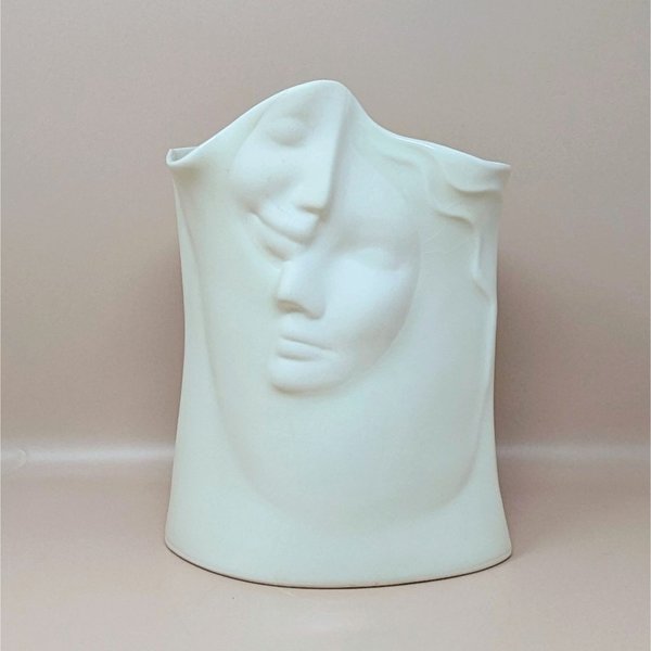 Front view of a white porcelain vase featuring two serene faces in relief. The faces overlap with soft, flowing lines against a neutral background.