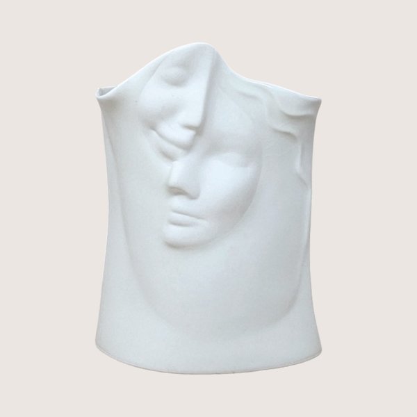 A matte white ceramic vase molded with two abstract interlocking faces, centered on a beige background.