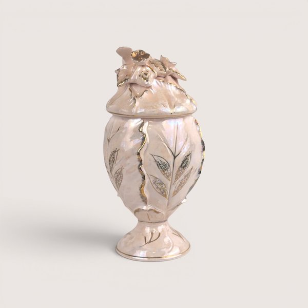 An iridescent pale pink ceramic compote jar with gold accents and a lid topped with a bird, fruit, and leaves.