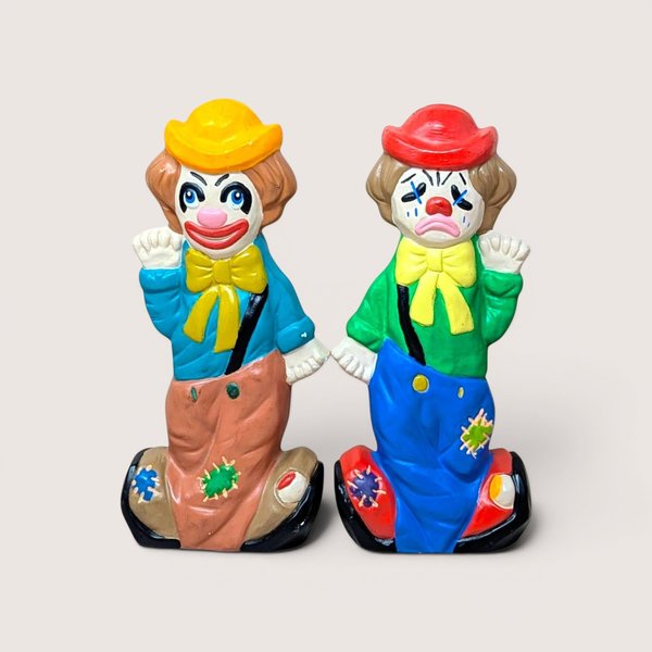 A pair of vintage ceramic hobo clown coin banks on a plain background, one smiling in a blue shirt and one crying in green.