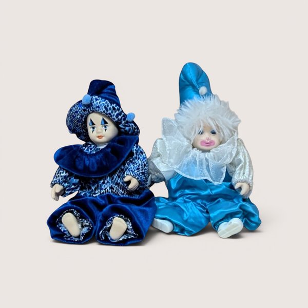 Two small vintage porcelain clown dolls sitting together in blue and white satin outfits, centered on a beige background.