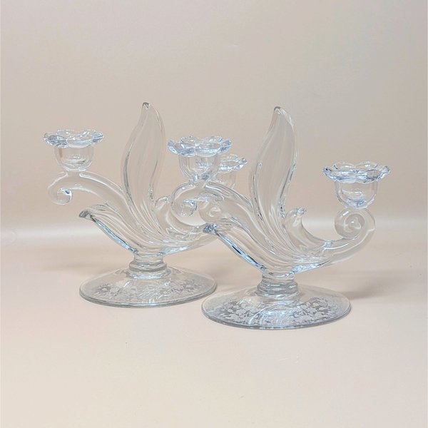 A pair of vintage clear glass double candlesticks by Duncan Miller in the First Love pattern. The holders feature elegant sweeping curves, flower-shaped candle cups, and an intricate etched floral design on the circular bases.