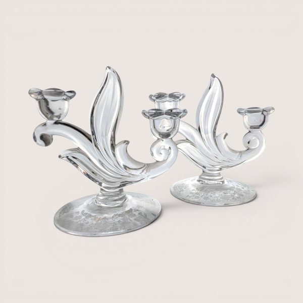A pair of elegant clear glass candelabras with curved leaf details and flower-shaped cups, centered on a beige background.