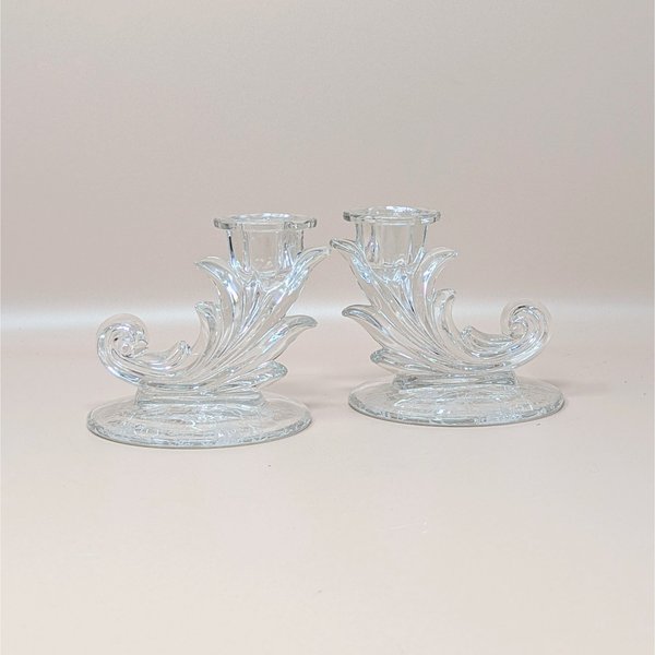 A pair of vintage clear glass cornucopia-shaped candlesticks by Fostoria in the Navarre pattern. The holders feature a decorative Baroque scroll design and an intricate etched floral pattern on the circular bases.