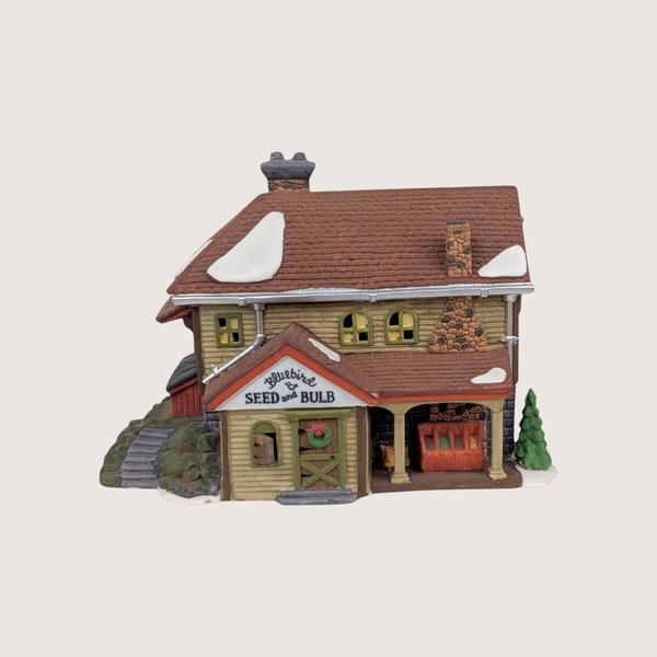 Department 56 Bluebird Seed and Bulb ceramic garden shop building with a stone chimney and snow on the roof.