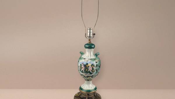 A tall Italian ceramic table lamp featuring a green and white urn body with hand-painted cherubs. The lamp sits on an ornate gold-colored metal base and has small holes in the ceramic for an internal glow.