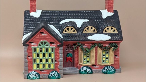 A large red brick ceramic miniature house with a snow-covered roof and two chimneys. It features festive green garlands and a wreath on the front door.