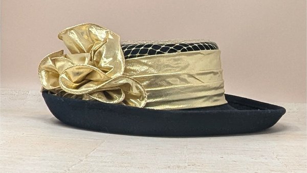 A black wool ladies' hat with a flat top and wide brim. It is decorated with a wide metallic gold band, a gold fabric rose, and black mesh netting on the crown.