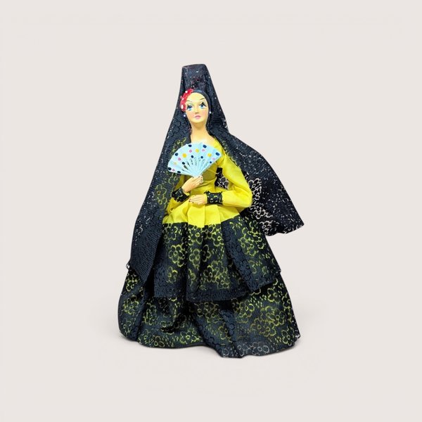 Vintage Spanish flamenco dancer doll wearing a yellow dress and black lace mantilla while holding a fan, centered on a plain linen background.