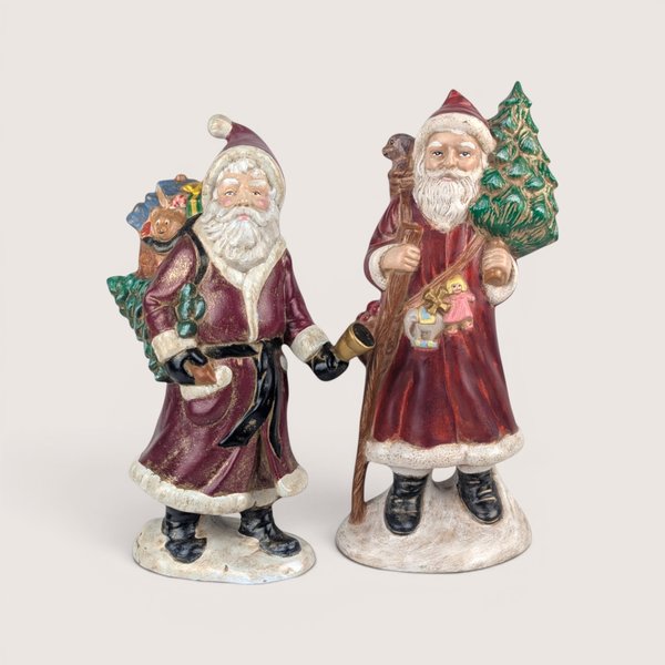 Pair of vintage Santa Claus figurines carrying Christmas trees and sacks of toys, centered on a plain linen background.