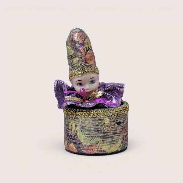 Vintage porcelain jester doll emerging from a matching round metallic fabric box with gold trim, centered on a plain linen background.