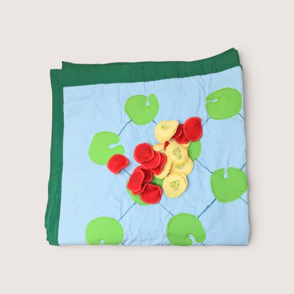 Folded fabric game mat featuring a light blue background patterned with green lily pads, topped with a pile of small red and yellow fabric tokens, centered on a plain linen background.