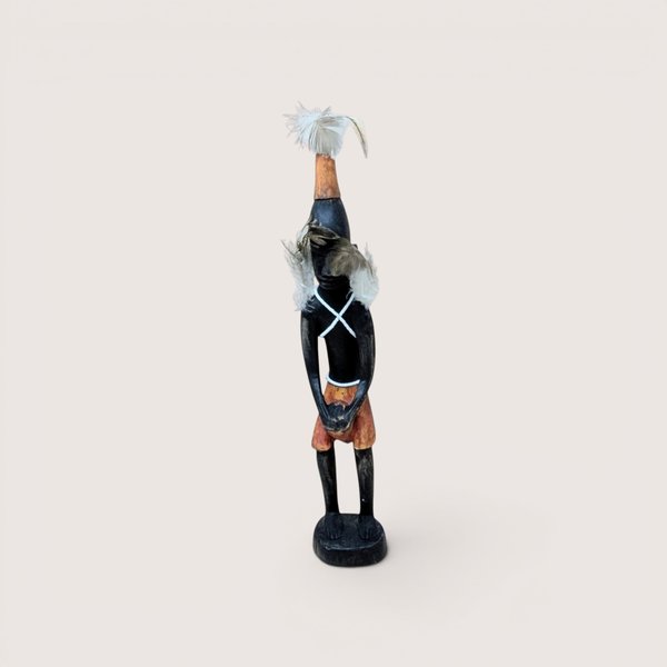 Carved wooden tribal figurine standing on a circular base, featuring feather details on the headpiece and shoulders with a white criss-cross chest pattern, centered on a plain linen background.