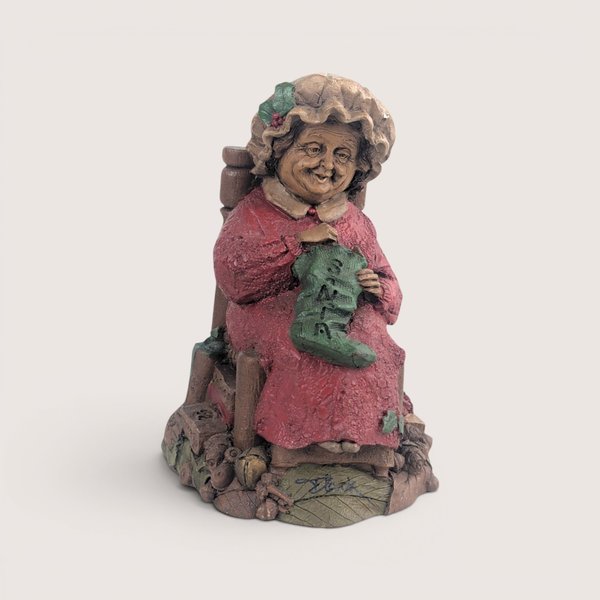 A vintage Tom Clark Cairn Studios resin figurine titled Mrs Claus featuring a woman sitting in a chair knitting a stocking.