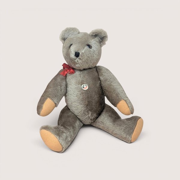 Vintage mohair teddy bear with grey-brown fur, a small red bow neck tie, tan felt paw pads, and a round chest tag, centered on a plain linen background.