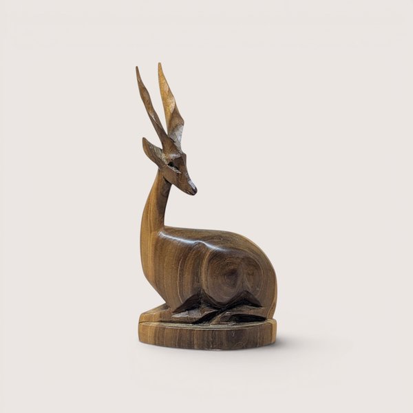 Hand-carved wooden gazelle or antelope figurine depicted in a recumbent, bedded position on an oval base, featuring prominent twisted horns and a rich, polished natural wood grain finish, centered on a plain linen background.