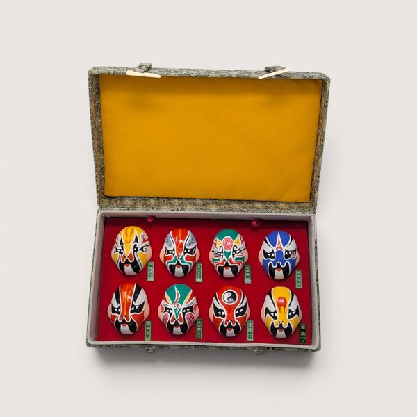 Boxed set of eight vintage miniature Chinese opera masks, hand-painted with vibrant traditional designs and arranged in two rows of four inside a red-lined display case with a bright yellow fabric lid interior, centered on a plain linen background.