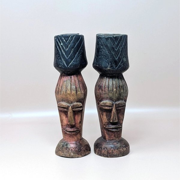 A pair of vintage hand carved wooden candlestick holders on a soft pink background. The dark brown pillars feature stylized tribal or tiki faces topped with dark, chevron carved candle cups and flare out into circular bases.