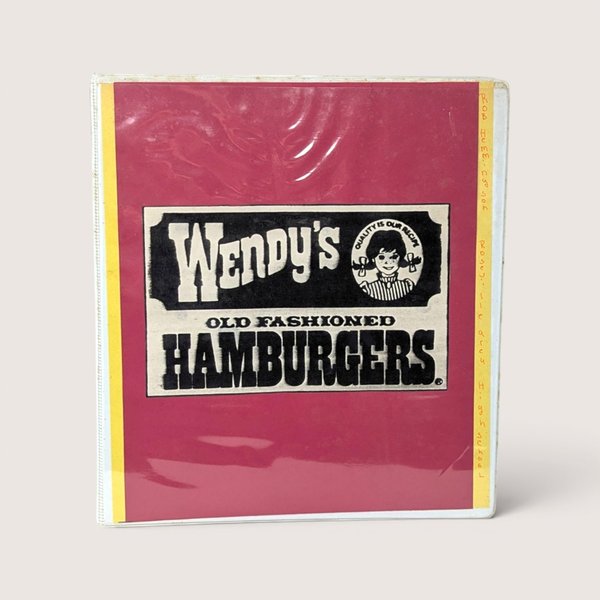 A vintage 1980s Wendy's employee instruction manual in a red three ring binder on a linen background