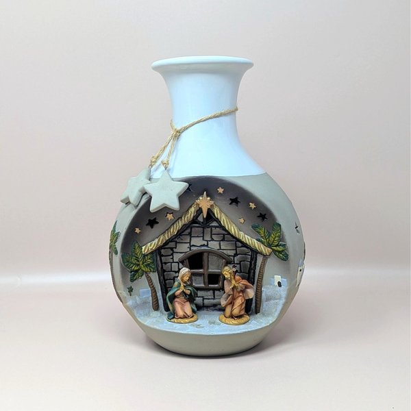A vintage cream and gray Roman Inc ceramic light up nativity vase on a soft pink background. The round vase features a molded diorama window showing Mary and Joseph at the stable beneath cut out star openings and a hanging star charm.