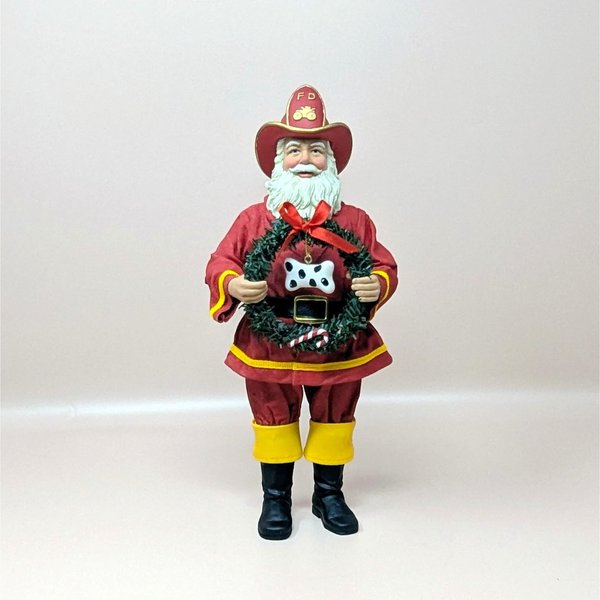 A vintage Fabriche fireman Santa Claus figurine on a soft pink background. Standing eleven and a half inches tall, the figure wears a red firefighter uniform and hat while holding a green faux pine wreath adorned with a spotted dog bone and red ribbon.