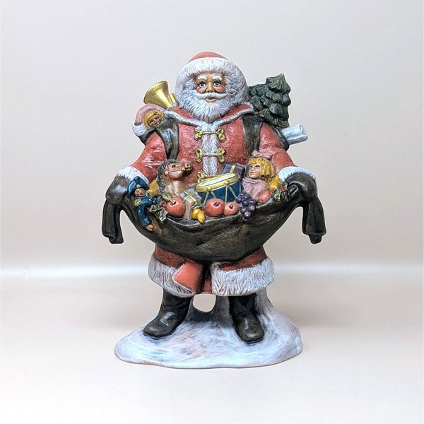 A vintage hand painted ceramic Santa Claus figurine on a soft pink background. Standing nine and a half inches tall, the classic robed Santa holds open a massive black sack overflowing with colorful toys including a drum, doll, and horse.