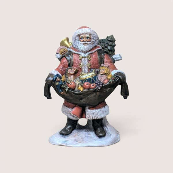 Vintage standing Santa Claus figurine dressed in a classic red, fur-trimmed coat and black boots, holding out a large overflowing sack filled with traditional toys including a drum, a rocking horse, a doll, a toy soldier, a brass horn, and wrapped gifts, standing on a snow-covered base and centered on a plain linen background.