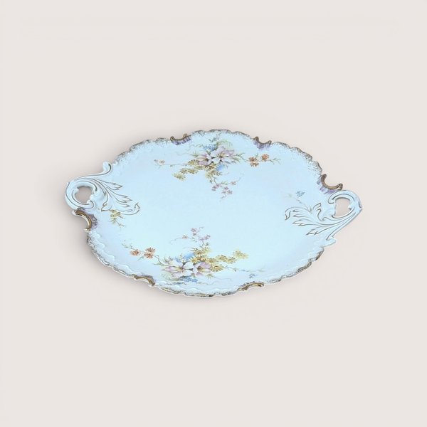 Vintage porcelain serving platter or cake plate with an elegant ruffled rim and twin integrated scroll handles, decorated with delicate hand-painted pastel floral sprays on a pale blue background with gold-gilt highlights, centered on a plain linen background.