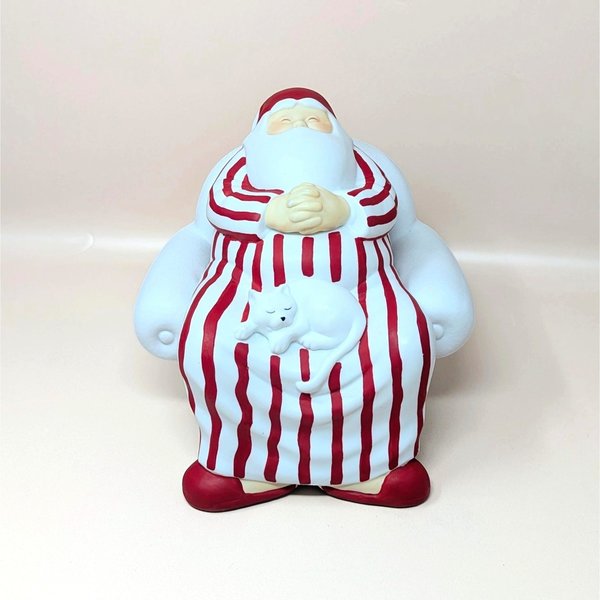 A vintage Department 56 Winter Silhouettes "Cat Nap" Santa figurine on a soft pink background. The matte white porcelain sculpture shows Santa sleeping soundly in a large armchair wearing red-striped pajamas with a white cat napping on his lap.