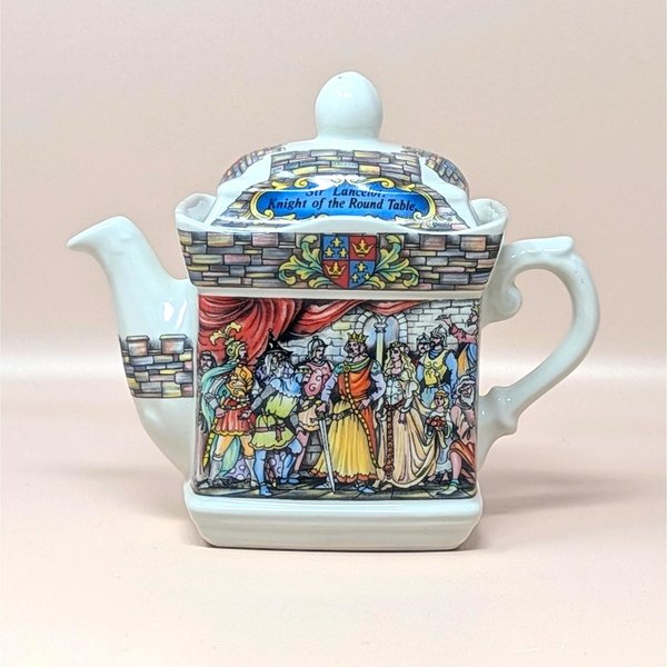 A vintage Sadler porcelain castle themed teapot on a soft pink background. The square cream colored teapot features a detailed double sided illustration of King Arthur's court at Camelot and Sir Lancelot with stone wall borders and a matching lid.