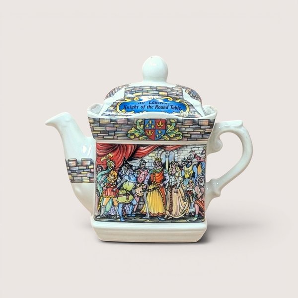 A vintage Sadler porcelain castle themed teapot on a soft pink background. The square cream colored teapot features a detailed double sided illustration of King Arthur's court at Camelot and Sir Lancelot with stone wall borders and a matching lid.