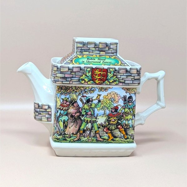 A vintage Sadler porcelain castle themed teapot on a soft pink background. The square cream colored teapot features a detailed double sided illustration of the Sheriff of Nottingham at his table and Robin Hood in Sherwood Forest with stone walls.