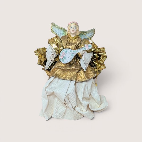 A vintage paper mache and fabric angel tree ornament on a soft pink background. The nine inch tall angel features a metallic gold upper gown, stiffened cream fabric crinkle skirt, soft gold wings, and holds a small blue stringed lute.