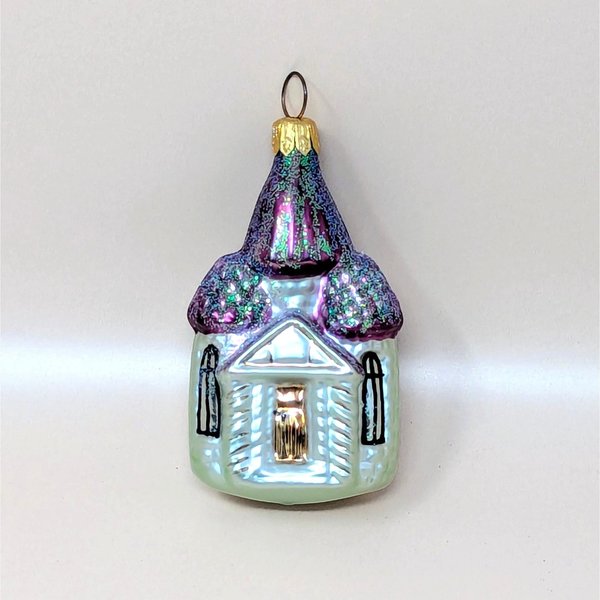A vintage Christopher Radko blown glass church ornament with its box on a soft pink background. The fine mercury glass cathedral features iridescent pale green walls, hand painted black windows, and shimmering purple steeples covered in sparkling glitter.