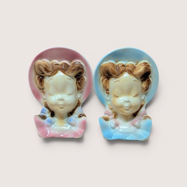Two vintage ceramic lady head wall pocket planters wearing pink and blue bonnets side by side on a plain background