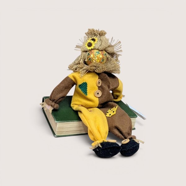 A vintage fabric scarecrow shelf sitter with a burlap hat and patterned face sitting on a green book against a plain background