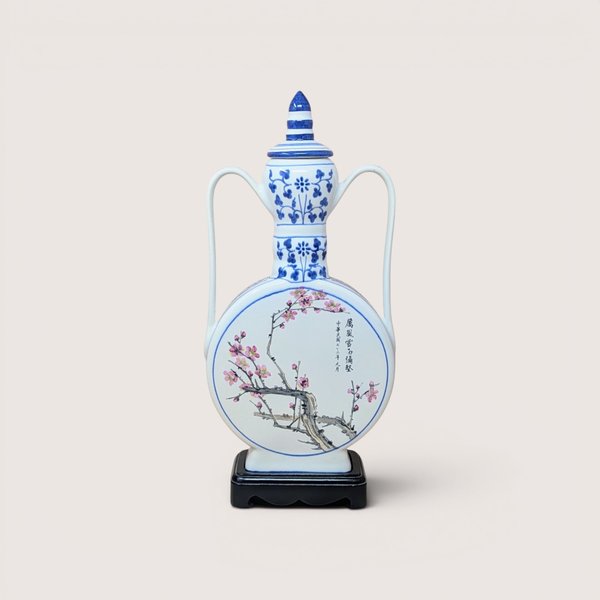 A large blue and white porcelain moon flask with pink plum blossoms and Chinese calligraphy on a wooden stand against a plain background