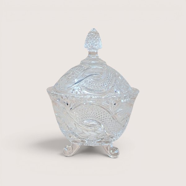 A vintage clear crystal candy dish with a matching lid and three scroll feet on a plain background