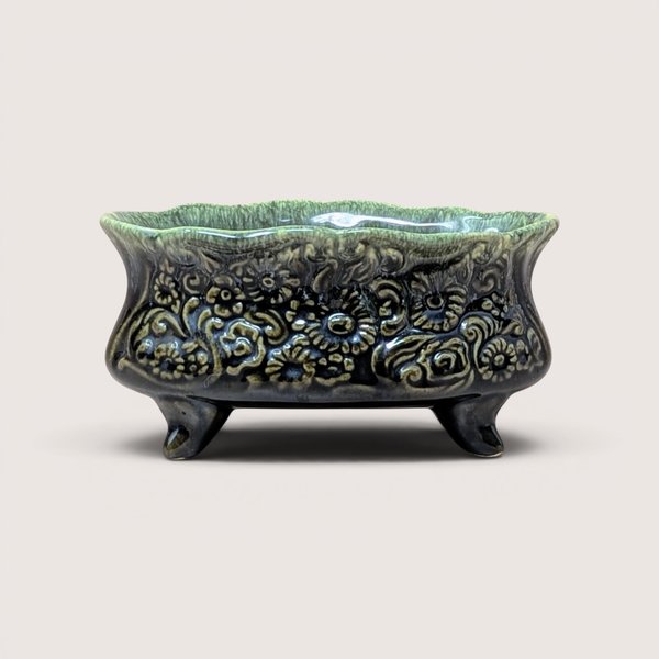 A vintage footed green ceramic planter with detailed embossed flowers resting on a plain beige background