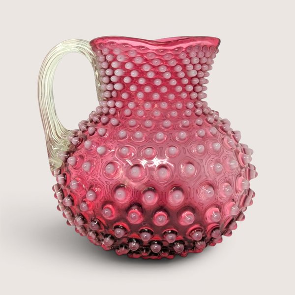 A vintage cranberry glass pitcher with an applied clear handle and an allover raised hobnail pattern on a plain background