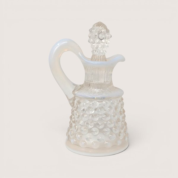 A vintage Fenton white opalescent hobnail glass cruet with a matching stopper and handle on a light cream background